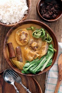 a wooden bowl filled with food next to rice and spoons on top of a table