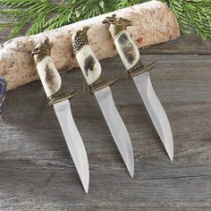 three knives are sitting next to each other on a piece of wood with pine branches in the background