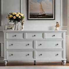 a white dresser with flowers on top and a painting hanging above it in a living room