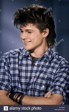 a young man with his arms crossed smiles at the camera while wearing a plaid shirt