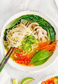 two bowls filled with noodles and vegetables on top of a white table next to chopsticks