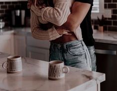 a man and woman hugging in the kitchen with two mugs on the counter top