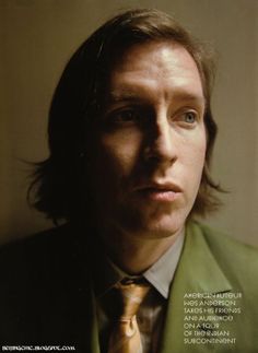 a man with long hair wearing a green suit and gold tie, looking at the camera