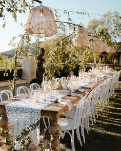 an outdoor dining table set up with white chairs and tables covered in lace, surrounded by greenery