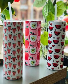 three cups with apples and watermelon designs are sitting on a window sill