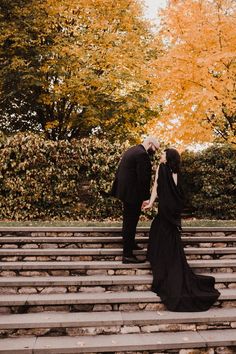 a man and woman kissing on the steps in front of trees with leaves turning yellow