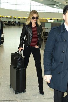 a woman in black jacket and sunglasses pulling luggage behind her at an airport with another man