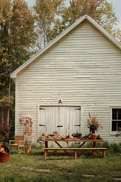 an old white barn with a picnic table and chairs in front of it, surrounded by fall foliage