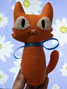 a hand holding a small orange cat with big eyes and a blue ribbon around its neck