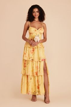 a woman wearing a yellow dress with flowers on the bottom and side, standing in front of a beige background