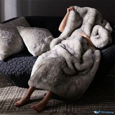 a woman laying on top of a black couch covered in grey furry blankets and pillows