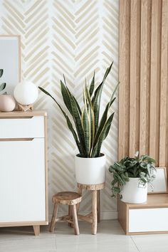 a plant in a white pot next to a wooden stool and wall with geometric pattern