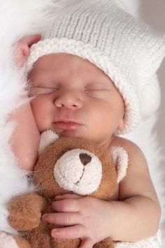 a baby sleeping with a teddy bear in his arms and wearing a white knitted hat