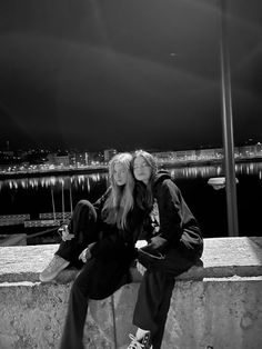 two young women sitting on the edge of a concrete wall at night with city lights in the background