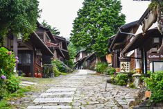 a cobblestone street lined with wooden buildings and lots of greenery on both sides