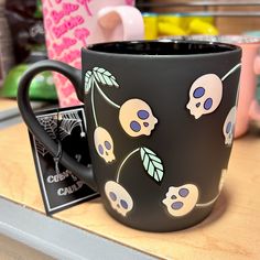 a black coffee mug with skulls and leaves on it sitting on a counter next to other items