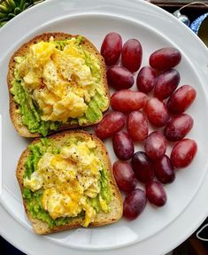 two pieces of toast with eggs, avocado and grapes on it sitting on a white plate