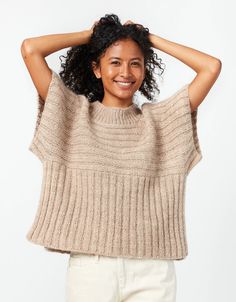 a smiling woman with her hands on her head, wearing a beige sweater and white pants