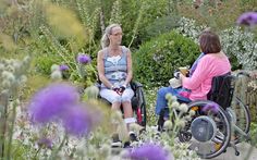 two women in wheelchairs talking to each other on a path surrounded by wildflowers