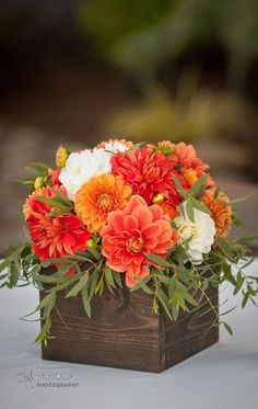 an arrangement of orange and white flowers in a wooden box on a table with greenery