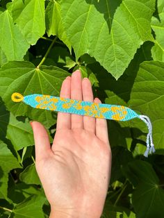 a hand is holding a blue and yellow beaded object in front of green leaves