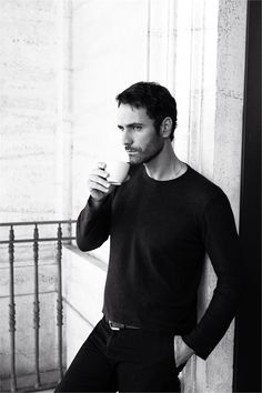 a man leaning against a wall holding a coffee cup in one hand and looking at the camera