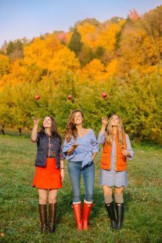 three young women are standing in the grass and one is reaching up for an apple