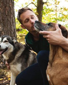 a man sitting next to two dogs in the woods with their noses close to each other