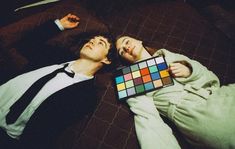 two people laying on a bed with one holding a color swatter and the other looking up