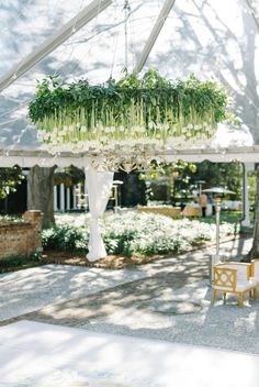 a white tent with chairs and plants hanging from it's ceiling in the middle of a garden