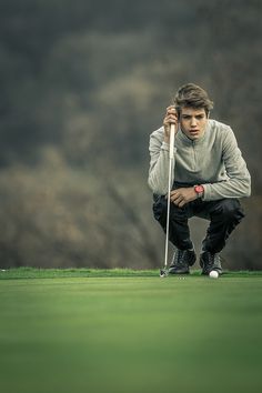 a young man kneeling down on top of a green golf field holding a white club