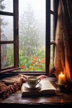 an open book and cup on a table in front of a window with rain coming down