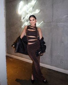 a woman standing in front of a neon sign wearing a black top and brown skirt