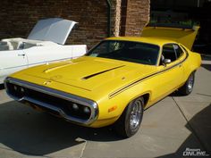 a yellow muscle car parked in front of a white boat on the water next to a brick building