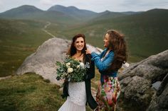 two beautiful women standing next to each other on top of a mountain holding bouquets