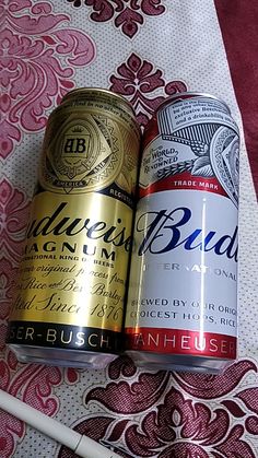 two cans of beer sitting next to each other on a tablecloth with a pen