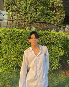 a young man standing in front of a bush wearing a green hat and white shirt