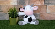 a stuffed cow sits next to a potted plant on the grass in front of a brick wall