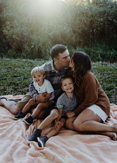 a family is sitting on a blanket in the grass and kissing each other while they are surrounded by greenery