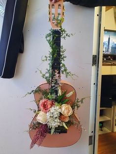 a guitar hanging from the side of a wall with flowers and greenery on it