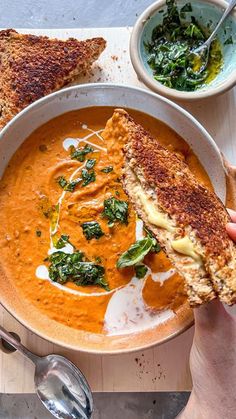 a person holding a piece of bread over a bowl of red soup with spinach on the side