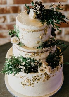 a three tiered wedding cake with flowers and greenery on the top, sitting on a table in front of a brick wall