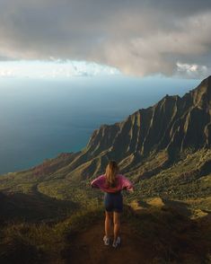 a woman standing on top of a lush green hillside next to the ocean under a cloudy sky