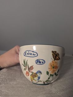 a hand holding a bowl with flowers and birds painted on the side, in front of a gray wall