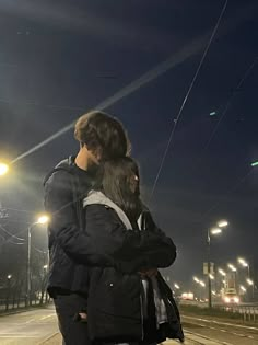 a man and woman standing next to each other on a sidewalk at night with street lights in the background