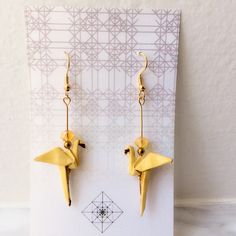 a pair of yellow bird shaped earrings sitting on top of a white card with an origami background