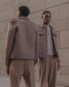 two men standing next to each other in front of a wall wearing jackets and slacks