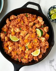 a skillet filled with rice and chicken next to limes on a table top