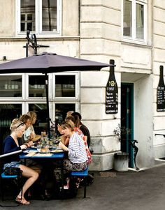 three women are sitting at a table outside with an umbrella over their heads and one woman is eating