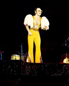 a man standing on top of a stage next to another man in yellow pants and white wings